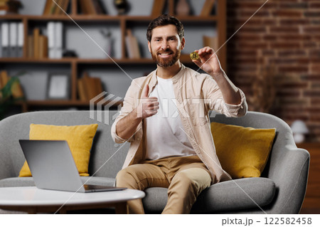 A man with a beard is happily holding up a cookie while seated on a comfortable sofa in a stylish living room. The laptop in front of him suggests he is enjoying a relaxing break. 122582458