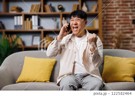 hear good news, talking phone, Excited man, An enthusiastic man sits on a couch, joyfully reacting while speaking on the phone. Yellow cushions add warmth to the stylish living room filled with books 122582464