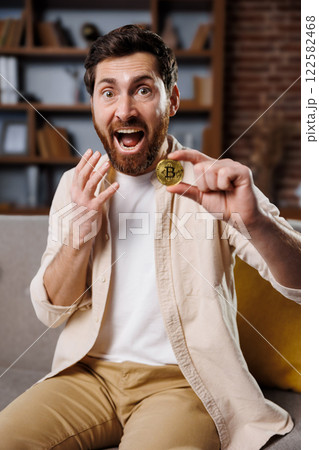 excited man, shows cryptocurrency, gold bitcoin coin, man with a beard and cheerful expression holds up a bitcoin in a well-decorated living room. His excitement reflects the growing interest in 122582468