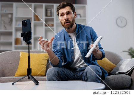 online broadcast, lesson, video call, man actively participates in a virtual meeting, gesturing with his hands. He holds a tablet and is positioned on a comfortable couch in a contemporary living room online broadcast, lesson, video call, man actively participates in a virtual meeting, gesturing with his hands. He holds a tablet and is positioned on a comfortable couch in a contemporary living room 122582502