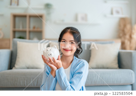 Young Asian woman holding piggy bank and sitting on the floor at home 122583504