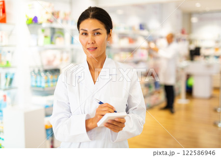 Positive female pharmacist makes notes on a notebook in the sales area of pharmacy 122586946