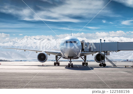 Front view of the white wide body passenger aircraft at the airbridge on the airport apron on the background of high snow capped mountains Front view of the white wide body passenger aircraft at the airbridge on the airport apron on the background of high snow capped mountains 122588156