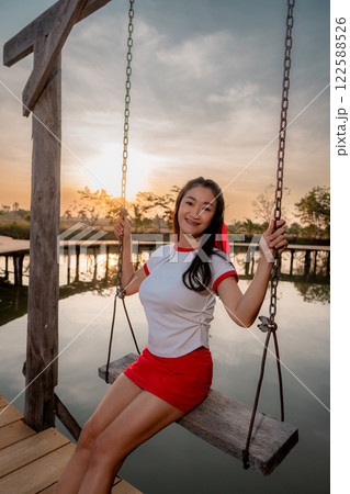 Asian woman poses cutely, sitting on a swing, on a wooden bridge, backlit photo. 122588526