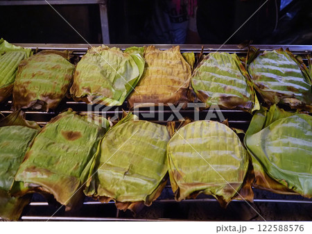 A Grilled banana leaf wrapped snacks on barbecue grill, showcasing traditional street food preparation. leaves are charred, indicating smoky flavor 122588576