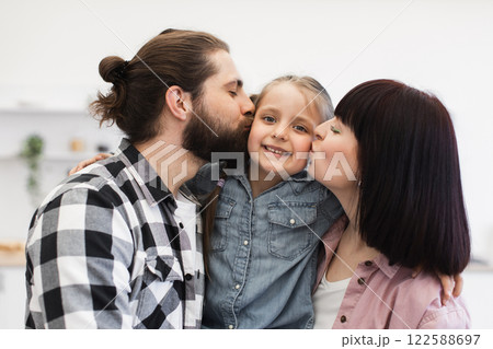 Caucasian mom and dad lovingly kiss young daughter in bright kitchen setting, showcasing joy and togetherness. 122588697