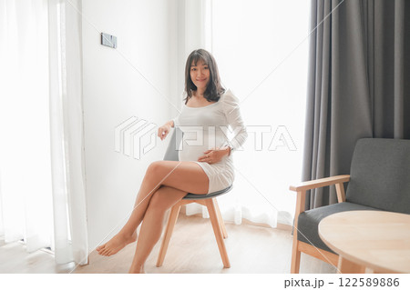 Pregnant Asian woman in white dress in bedroom Pregnant Asian woman in white dress in bedroom 122589886