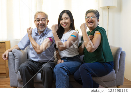 Senior Asian couple and teen woman get vaccinated with bandage on arm show thumb up sign in living room. Grandfather and grandmother get vaccine. 122590158