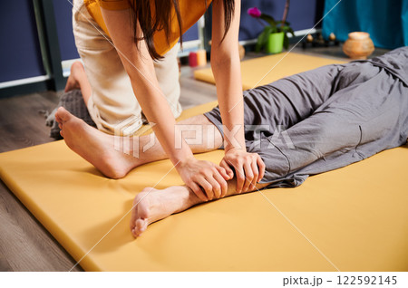 Masseuse making thai yoga massage. Cropped view of woman therapist doing traditional massage treatment for male patient. Client lying on mat, meditating, enjoying hand massage in calm atmosphere. Masseuse making thai yoga massage. Cropped view of woman therapist doing traditional massage treatment for male patient. Client lying on mat, meditating, enjoying hand massage in calm atmosphere. 122592145