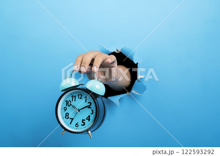 Human hand holding alarm clock through ripped paper. Mockup or Copy space. Concept of time management, reminder or planning, deadline, timetable and countdown 122593292