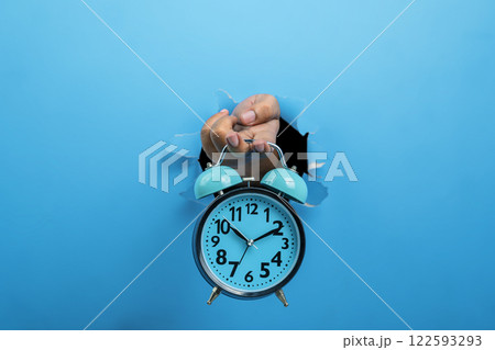 Human hand holding alarm clock through ripped paper. Mockup or Copy space. Concept of time management, reminder or planning, deadline, timetable and countdown Human hand holding alarm clock through ripped paper. Mockup or Copy space. Concept of time management, reminder or planning, deadline, timetable and countdown 122593293