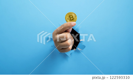 A human hand is holding a coin of bitcoin through ripped paper. Mockup or Copy space. Concept of cryptocurrency, economy, finance, and investment A human hand is holding a coin of bitcoin through ripped paper. Mockup or Copy space. Concept of cryptocurrency, economy, finance, and investment 122593310