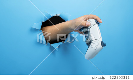 Human hand holding the gamepad, joystick, or video game controller through ripped paper. mockup or copy space. Concept of playing video games, entertainment or leisure activity Human hand holding the gamepad, joystick, or video game controller through ripped paper. mockup or copy space. Concept of playing video games, entertainment or leisure activity 122593311
