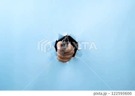 Human hand holding handgun through ripped paper. Hand gestures. Mockup or Copy space. Aiming weapons, war, crime, or violence 122593600