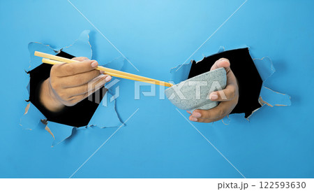 A human hand is holding a wooden chopstick and bowl through ripped paper. Hand gestures to reach or grab food. Mockup or Copy space A human hand is holding a wooden chopstick and bowl through ripped paper. Hand gestures to reach or grab food. Mockup or Copy space 122593630