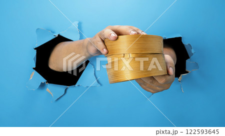 A human hand is holding a bamboo steamer through ripped paper. Hand gestures to serve food. Mockup or Copy space A human hand is holding a bamboo steamer through ripped paper. Hand gestures to serve food. Mockup or Copy space 122593645