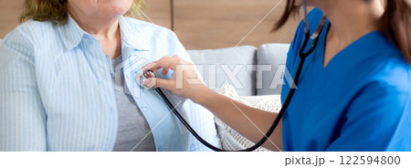 Caregiver young nurse woman examining elderly with stethoscope for checkup sitting on sofa. 122594800