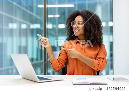 Businesswoman at workplace inside office, pointing with hands and fingers to the side, joyful office worker, recommends approvingly. Woman working with laptop at workplace near window. 122594963