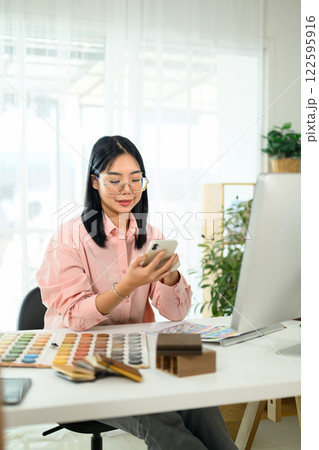 Young creative woman sitting at modern workspace checking project updates on smartphone 122595916