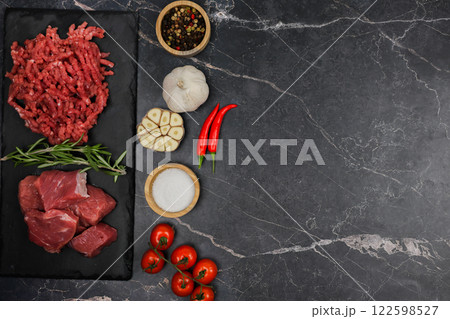 Top view of chunks of beef lying on a black cutting board, separated by sprigs of rosemary with red minced meat, vegetables and seasonings on a dark background with space for text. The concept of Top view of chunks of beef lying on a black cutting board, separated by sprigs of rosemary with red minced meat, vegetables and seasonings on a dark background with space for text. The concept of 122598527