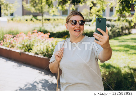 Hi. Female in sunglasses taking a selfie photo using her mobile phone in the public park. Woman in t-shirt recording video, talking on online call and relaxing outdoors. Urban people concept. Hi. Female in sunglasses taking a selfie photo using her mobile phone in the public park. Woman in t-shirt recording video, talking on online call and relaxing outdoors. Urban people concept. 122598952