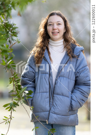 Young woman wearing blue puffer jacket is posing near green leaves 122600011