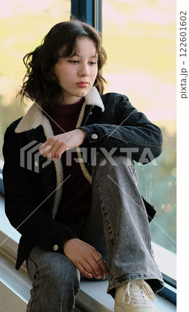 A contemplative girl sits by a window, her gaze directed outward. The photo highlights a reflective mood, symbolizing personal growth and the beauty of stillness. 122601602