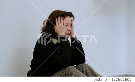 A girl with a solemn expression rests her head in her hand, seated in a minimalistic space. This photo represents feelings of loneliness, worry, or personal reflection. 122601605