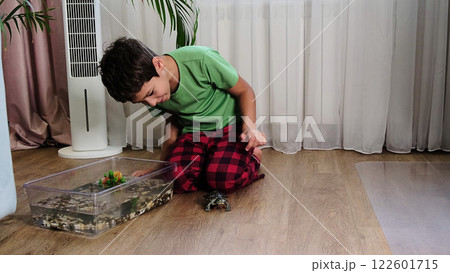 young boy kneels next to his turtle, which is on the floor near its habitat tank filled with water and aquatic decor. The image reflects the curiosity and care that children can have for their pets. 122601715