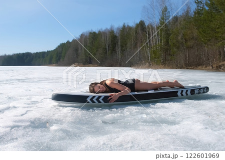 Woman in swimsuit basking in sun lying on sup board on frozen ice on river lake. Woman in swimsuit basking in sun lying on sup board on frozen ice on river lake. 122601969