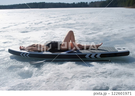 Woman in swimsuit basking in sun lying on sup board on frozen ice on river lake. 122601971