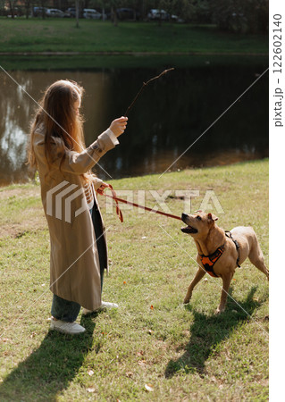 young woman walks with her dog in the park. A lady in a beige raincoat is playing with a pit bull young woman walks with her dog in the park. A lady in a beige raincoat is playing with a pit bull 122602140