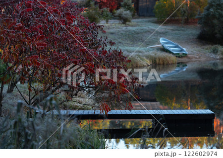 Autumnal landscape with a boat, wooden footbridge and red leaved bush on the shore of a lake in Latvia 122602974
