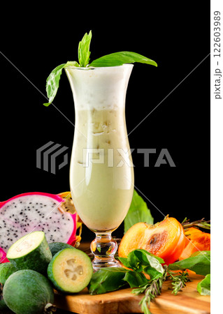Glass of fresh fruit smoothie decorated with basil, feijoa, persimmon and dragon fruit at wooden table isolated at black background. 122603989