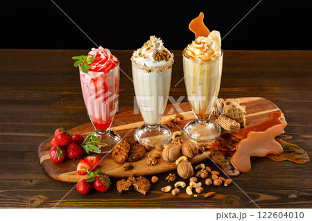 Three sweet milkshakes with nuts, caramel, strawberry and whipped cream at a wooden board on table background. 122604010