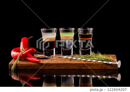 Three alcohol shooter layered cocktails on a wooden board served with barman spoon and chili pepper isolated at black background. Three alcohol shooter layered cocktails on a wooden board served with barman spoon and chili pepper isolated at black background. 122604207
