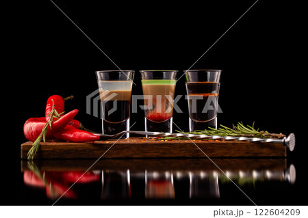 Three alcohol shooter layered cocktails on a wooden board served with barman spoon and chili pepper isolated at black background. Three alcohol shooter layered cocktails on a wooden board served with barman spoon and chili pepper isolated at black background. 122604209