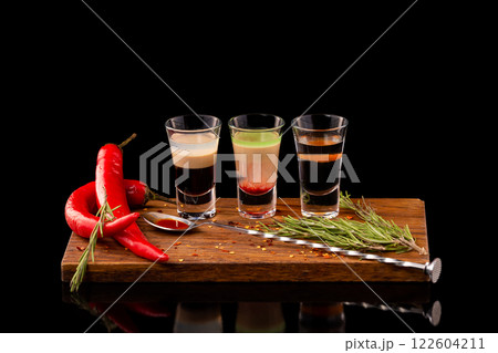 Three alcohol shooter layered cocktails on a wooden board served with barman spoon and chili pepper isolated at black background. Three alcohol shooter layered cocktails on a wooden board served with barman spoon and chili pepper isolated at black background. 122604211