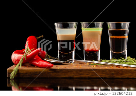 Three alcohol shooter layered cocktails on a wooden board served with barman spoon and chili pepper isolated at black background. 122604212