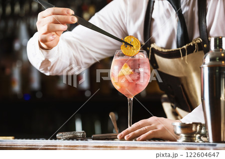 Elegant bartender is preparing pink cocktail holding orange chips at bar counter background. Elegant bartender is preparing pink cocktail holding orange chips at bar counter background. 122604647