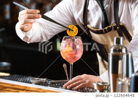 Elegant barman is making pink cocktail holding orange chips at bar counter background. 122604648
