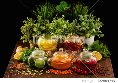 Set of colorful glass tea pots decorated with green plants and berries on a wooden table board at dark background. 122604743
