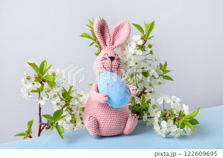 Knitted toy pink bunny with blue Easter egg and with cherry blossoms on blue table and gray background. Concept of spring, Easter Knitted toy pink bunny with blue Easter egg and with cherry blossoms on blue table and gray background. Concept of spring, Easter 122609695