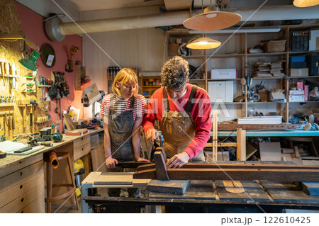 Beginner woman watch master carpenter operate saw machine in workshop. Husband and wife collaborate 122610425