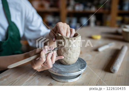 Hands of woman sculpting with loop tool, carving pattern on clay mug while removing excess material Hands of woman sculpting with loop tool, carving pattern on clay mug while removing excess material 122610435