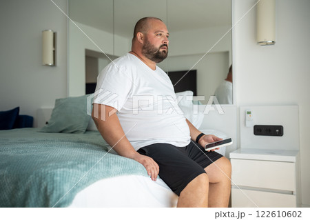 Lazy overweight man sits on hotel bed with phone, gaze thoughtfully at one point. Loneliness, fear Lazy overweight man sits on hotel bed with phone, gaze thoughtfully at one point. Loneliness, fear 122610602