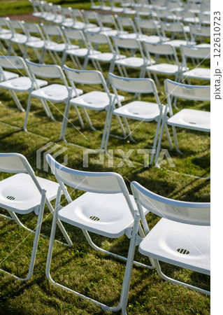 Folding chairs standing in row on green grass, casting shadow on ground. Outdoor party preparation 122610723