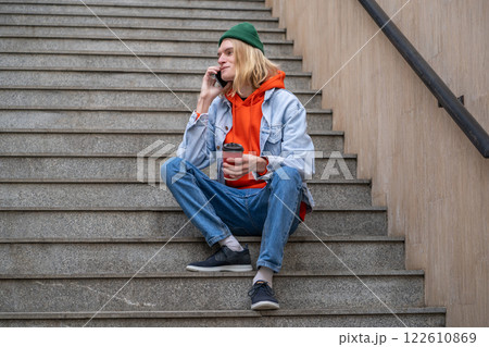 Pleased hipster guy sits on stairs outside smiling speak on cellphone holding takeout cup of coffee. 122610869
