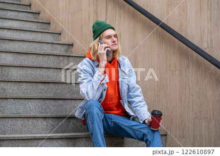 Hipster guy with coffee cup in hand sits on steps talks on smartphone in city street.  122610897