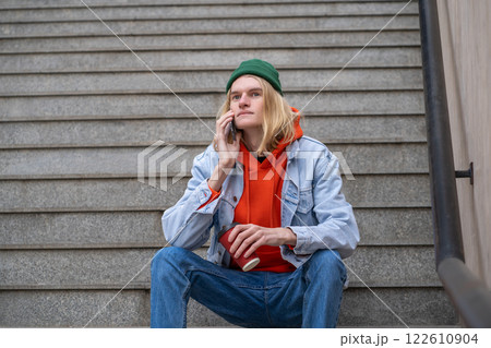 Hipster blond guy with coffee cup in hand sits on steps talks on smartphone in city street.  122610904